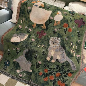 May include: Green woven throw blanket featuring whimsical illustrations of a goose, duck, cats, and flowers. The blanket has a fringed edge and a repeating pattern of a garden scene with orange trees and floral accents.
