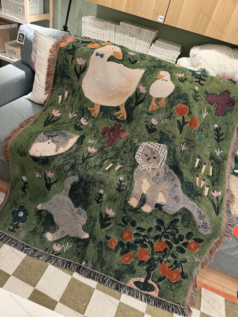 May include: Green woven throw blanket featuring whimsical illustrations of a goose, duck, cats, and flowers. The blanket has a fringed edge and a repeating pattern of a garden scene with orange trees and floral accents.