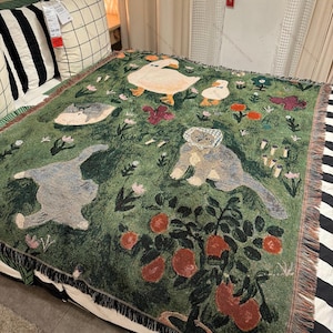 May include: Green woven throw blanket featuring whimsical illustrations of ducks, cats, and a bear. The blanket has a fringe border and a pattern of flowers and fruit. The throw is placed on a bed with striped and checkered pillows.