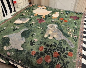 May include: Green woven throw blanket featuring whimsical illustrations of ducks, cats, and a bear. The blanket has a fringe border and a pattern of flowers and fruit. The throw is placed on a bed with striped and checkered pillows.