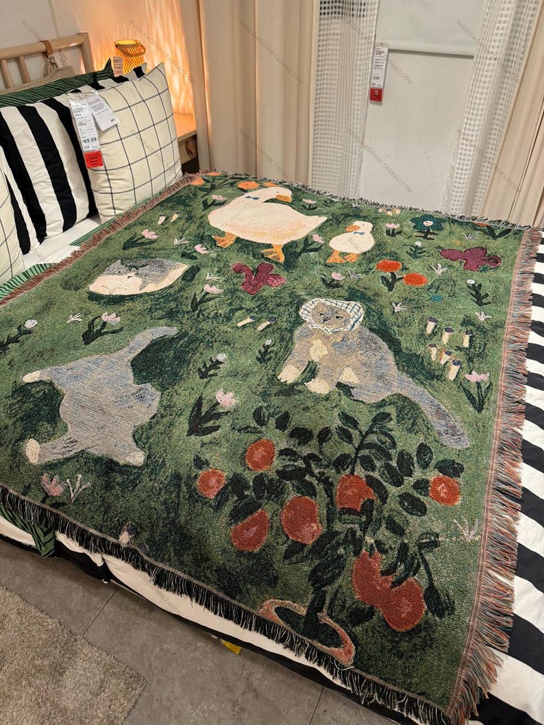 May include: Green woven throw blanket featuring whimsical illustrations of ducks, cats, and a bear. The blanket has a fringe border and a pattern of flowers and fruit. The throw is placed on a bed with striped and checkered pillows.