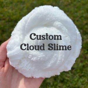 May include: A close-up of white cloud slime with the text "Custom Cloud Slime" in black. The fluffy, textured slime is held in a hand, with a blurred green background.