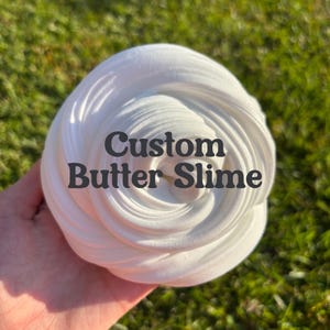 May include: A close-up of a white butter slime, swirled into a spiral shape. The text "Custom Butter Slime" is printed on the slime in a dark grey font. The background is blurred green grass.