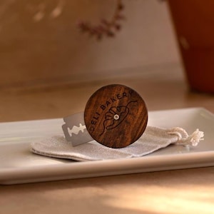 Op de afbeelding: Een houten scheermeshandvat met de tekst "DELI BAKERY" en een kleine afbeelding, samen met een metalen scheermesje. Het handvat ligt op een kleine stoffen zak, gepresenteerd op een witte rechthoekige plaat.