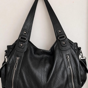 May include: A black shoulder bag featuring two vertical zipped pockets on the front, dual handles with metallic stud accents, and side compartments. The bag has a textured surface and silver-toned hardware, including zips and rectangular rings.