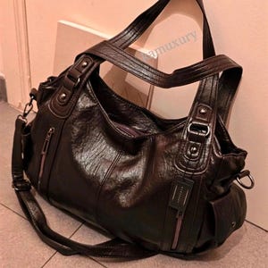 Puede incluir: Un bolso hobo de color marrón oscuro con acabado texturizado, que cuenta con dos asas superiores, una correa de hombro extraíble y bolsillos verticales con cremallera en la parte frontal. Incluye herrajes plateados y bolsillos laterales.