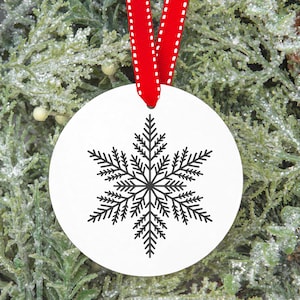 Op de afbeelding: Een witte, ronde kerstversiering met een zwart sneeuwvlokontwerp. De versiering hangt aan een rood lint met witte stiksels. De achtergrond is een groene, bevroren dennentak.