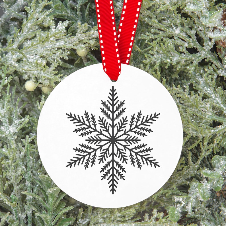 Op de afbeelding: Een witte, ronde kerstversiering met een zwart sneeuwvlokontwerp. De versiering hangt aan een rood lint met witte stiksels. De achtergrond is een groene, bevroren dennentak.