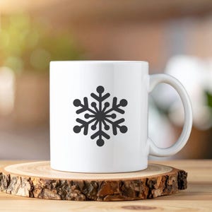 Op de afbeelding: Witte keramische mok met een zwart sneeuwvlokontwerp. De sneeuwvlok is gecentreerd en heeft een gedetailleerd, symmetrisch patroon. De mok heeft een gebogen oor en staat op een houten onderzetter.