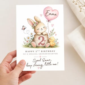 Könnte beinhalten: Eine Geburtstagskarte mit einem gezeichneten Hasen mit Blumenkranz, einem gelben Küken und einem rosa herzförmigen Luftballon mit der Aufschrift Emma. Ein Holzblock zeigt die Zahl 2. Text: Happy 2nd Birthday to a special granddaughter. Sweet Emma, keep shining, little one!