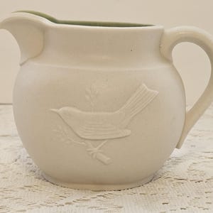 May include: Cream-colored ceramic pitcher with a bird relief design. The pitcher has a rounded body, a curved handle, and a spout. The interior rim is a light green color. The bird is perched on a branch.