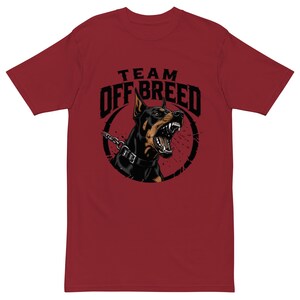Puede incluir: Una camiseta de manga corta color granate con un gráfico de un perro Doberman gruñendo que lleva un collar de pinchos y una cadena. El texto "TEAM OFF BREED" está impreso en negro sobre el perro, todo dentro de un marco circular negro.
