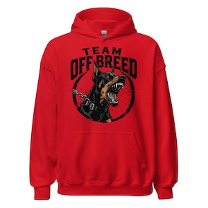 May include: A bright red hooded sweatshirt featuring a graphic print of a snarling Doberman dog wearing a spiked collar and chain. The text above the dog reads TEAM OFF BREED in bold black capital letters, all centered on the front of the garment.