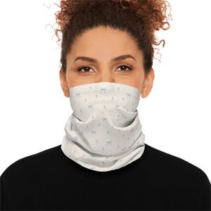 Könnte beinhalten: Ein weißes Halstuch mit einem sich wiederholenden Muster aus hellblauen Schleifen und kleinen floralen Akzenten. Das Halstuch wird um den Hals getragen und bedeckt das untere Gesicht. Die Person trägt ein schwarzes Oberteil.