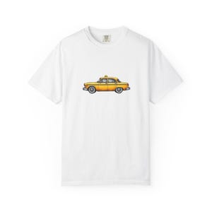 May include: White t-shirt featuring a yellow taxi cab illustration. The taxi has a checkered pattern on the side and a taxi sign on top. The car is a classic design, with chrome accents and white-walled tires.