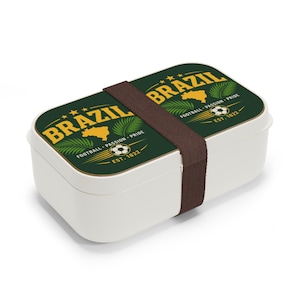 May include: A rectangular white lunch box secured with a brown elastic band. The lid features a dark green background with yellow text reading BRAZIL, a map of Brazil, palm leaves, a soccer ball, and the text FOOTBALL PASSION PRIDE EST. 1822.