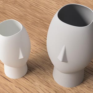 May include: Two white, head-shaped ceramic vases. The vases have a minimalist design, featuring a nose and an open top. One vase is smaller than the other. The vases are on a wooden surface.