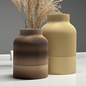 Puede incluir: Dos jarrones decorativos con detalles acanalados verticales. Uno es un degradado de tonos marrones, el otro es de color beige liso. Se coloca hierba ornamental seca en los jarrones. Los jarrones están sobre una superficie blanca.