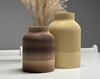 Puede incluir: Dos jarrones decorativos con detalles acanalados verticales. Uno es un degradado de tonos marrones, el otro es de color beige liso. Se coloca hierba ornamental seca en los jarrones. Los jarrones están sobre una superficie blanca.