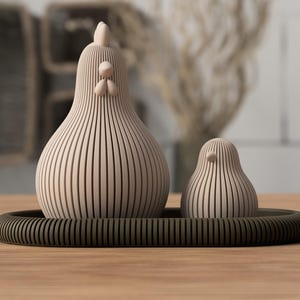 May include: A light beige chicken and a smaller chick figurine, both featuring a vertical ribbed texture, are positioned on a dark olive green oval tray. The chicken has a prominent comb and wattle, while the chick has a simple beak.