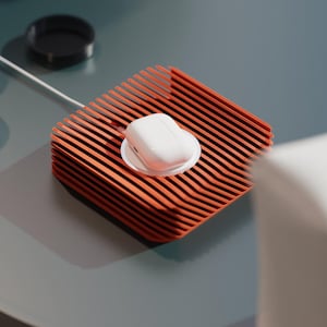 May include: An orange slatted tray with a white wireless charging case. The tray has a rectangular shape and a ridged design. The setup is on a grey surface, with a glass and a black ring in the background.