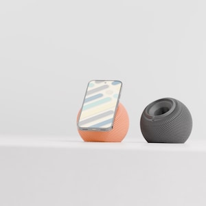 May include: A peach-coloured spherical phone stand holding a smartphone with a light blue and white abstract pattern on the screen, positioned next to a dark grey spherical phone stand with a circular indentation on the top surface.
