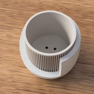 May include: A light gray, modern planter with a cylindrical interior and a textured, vertical-lined exterior. The planter has two drainage holes and is set on a wooden surface. The design is minimalist and suitable for small plants.