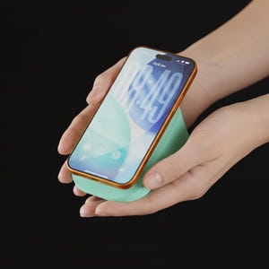 Peut inclure: Un support de téléphone vert menthe avec une bordure dorée, tenant un smartphone avec un écran bleu clair et blanc. Le téléphone affiche l'heure 09h40. Le support est tenu dans deux mains sur un fond noir.