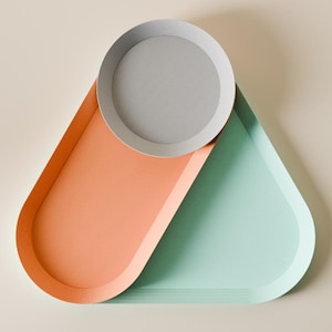 May include: A contemporary decorative tray set with a geometric design. The set features a round, light grey tray and two overlapping trays in coral and mint green. The trays are made of a smooth material and are set against a neutral background.