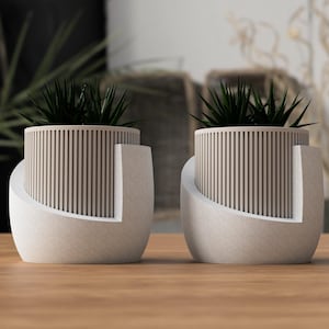 May include: Two modern planters with a light beige, vertical-slat design. The planters have a curved, off-white base and contain dark green plants. The planters are on a wooden surface.