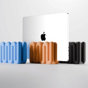 May include: A silver laptop with a black Apple logo is held upright by three wavy, U-shaped stands in blue, peach, and black. The stands feature a continuous, serpentine design that provides a stable base for the device on a flat surface.