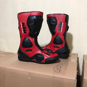 Könnte beinhalten: Paar rote und schwarze Motorradstiefel. Die Stiefel tragen das KTM Racing Team Logo und bestehen aus Leder und synthetischen Materialien. Sie haben verstärkte Bereiche zum Schutz und eine strapazierfähige Sohle.