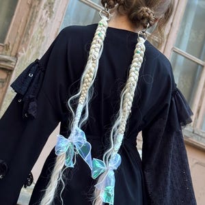 May include: A person with blonde braided hair, adorned with silver hoops and iridescent bows. The braids are long and hang down the back. The person is wearing a black dress with lace details on the sleeves. The background features a weathered wooden door.