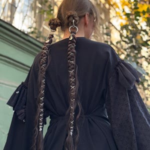 May include: A person's back is shown, wearing a black dress with ruffled sleeves. The hair is styled in two buns with silver rings and long braids adorned with silver accents. The background is blurred with green and yellow tones.