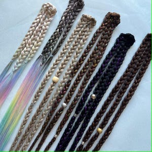 Op de afbeelding: Een verzameling gevlochten hairextensions in verschillende kleuren, waaronder blond, bruin en zwart. Sommige vlechten hebben kleurrijke, regenboogkleurige uiteinden. Verschillende vlechten zijn versierd met kralen en kleine metalen ringen.