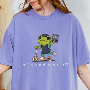 May include: A violet short-sleeved t-shirt featuring a graphic of a green frog wearing a dark blue scrub top, riding a pink scooter, and holding an x-ray film. The text below the frog reads, "off to do x-ray stuff" in white lowercase letters.