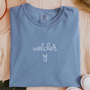 May include: A blue jean-colored t-shirt with the word "welder" in white script and a small bow. The shirt is laid flat with a hat, headband, scrunchie, shoes, and a leaf. The hat has a brown band.