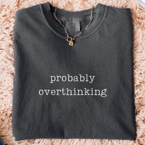 May include: A dark gray t-shirt with the phrase "probably overthinking" in white lettering. A gold chain necklace rests on the shirt. The shirt is on a peach-colored fluffy surface, with blue jeans and white sneakers visible.