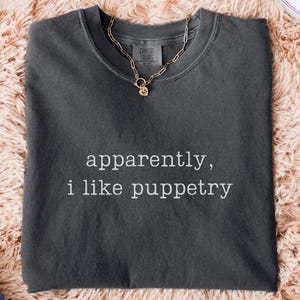 May include: A dark gray t-shirt with the text "apparently, i like puppetry" in white. A gold chain necklace with a pendant rests on the shirt. The shirt is on a peach-colored, fluffy surface, with blue jeans and white sneakers visible.