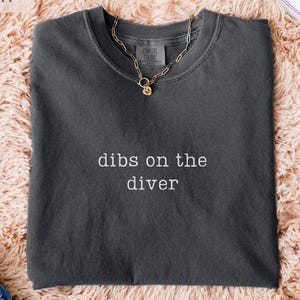 May include: A dark gray t-shirt with the text "dibs on the diver" in white. A gold chain necklace with a pendant is on the shirt. The shirt is on a peach-colored fluffy surface, with blue jeans and white sneakers visible.