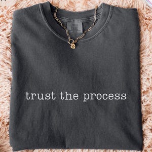 May include: A dark gray t-shirt with the phrase "trust the process" in white lettering. A gold chain necklace with a small pendant rests on the shirt. The shirt is on a peach-colored, fluffy surface, with a pair of white sneakers visible.