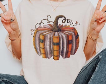 May include: An ivory t-shirt featuring a graphic of a pumpkin constructed from stacked, colorful vintage books with a textured stem. The shirt is worn by a model who is 5'4" and 120 lbs, wearing a size XL. The text IVORY is visible in the top corner.