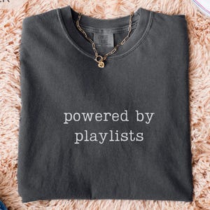 May include: A dark gray t-shirt with the text "powered by playlists" in white. A gold chain necklace with a pendant rests on the shirt. The shirt is folded on a peach-colored, fluffy surface, with a pair of blue jeans and white sneakers visible.