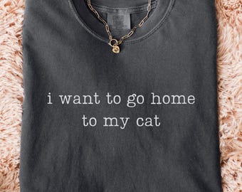 May include: A dark gray t-shirt with the text "i want to go home to my cat" in white. A gold chain necklace with a pendant is on the shirt. The shirt is on a fluffy, peach-colored surface, with blue jeans and white sneakers visible.