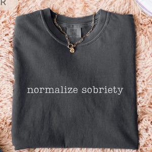 May include: A dark gray t-shirt with the phrase "normalize sobriety" printed in white. A gold chain necklace with a small pendant rests on the shirt. The shirt is displayed on a peach-colored, fluffy surface, with a pair of white sneakers and blue jeans visible.