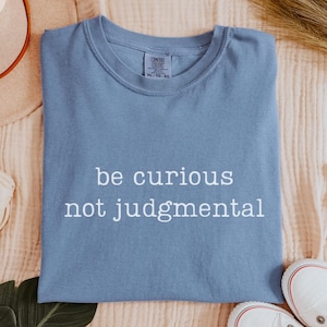 May include: A blue jean colored t-shirt with a crew neckline featuring the white text be curious not judgmental printed on the front. The shirt has a visible brand label at the inner neck area and is displayed flat on a textured fabric surface.