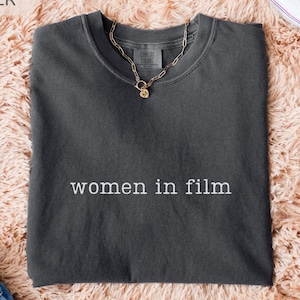 May include: A dark gray t-shirt with the text "women in film" in white. The shirt is laid flat on a peach-colored, fluffy surface. A gold chain necklace and white sneakers are also visible.