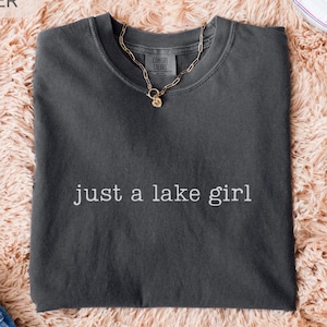 May include: A dark gray t-shirt with the text "just a lake girl" in white. A gold chain necklace with a pendant is on the shirt. The shirt is on a peach-colored fluffy surface, with blue jeans and white sneakers visible.