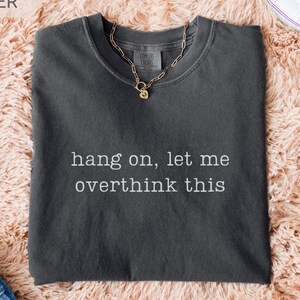 May include: A dark grey t-shirt with the slogan "hang on, let me overthink this" in white. A gold chain necklace with a small pendant is on the shirt. The shirt is on a pink fluffy surface.