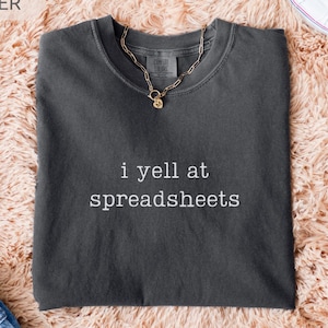 May include: Dark gray t-shirt with the phrase "i yell at spreadsheets" in white. A gold chain necklace and white sneakers are also visible. The shirt is laid on a peach-colored, fluffy surface.